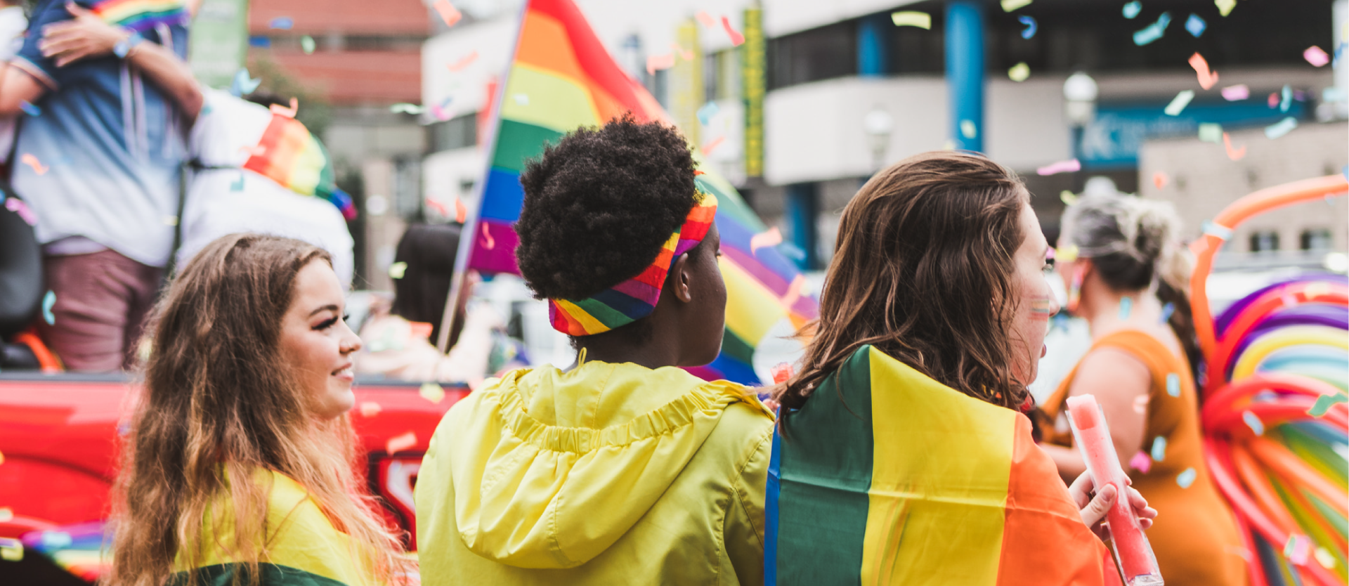 Three people watch the 2019 Pride Parade as it passes the Kingsplace mall. Two individuals are wearing rainbow flags as capes. The middle spectator is wearing a rainbow bandana as a headband. There is colourful confetti floating in the air. The float and walkers shown, from Saint Mary's First Nation, are decked out with rainbows and balloons. 