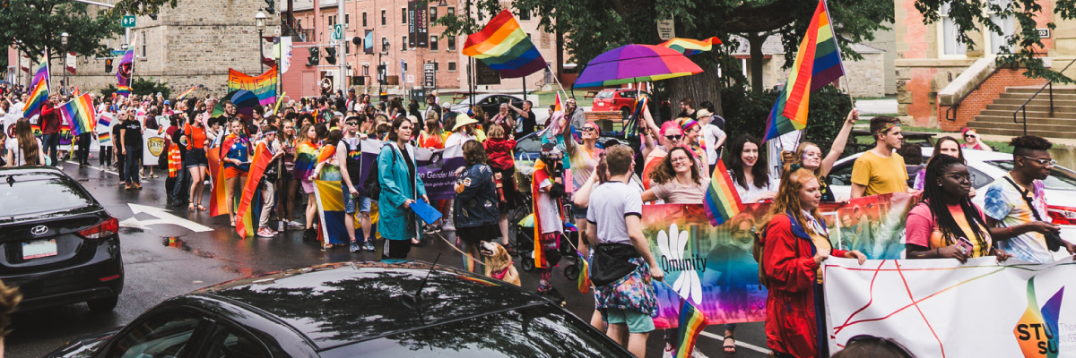An image of a parade, taken on Queen Street in Fredericton in 2019 by Tri Trinh. The photo shows many people with rainbow flags and banners in colourful attire.